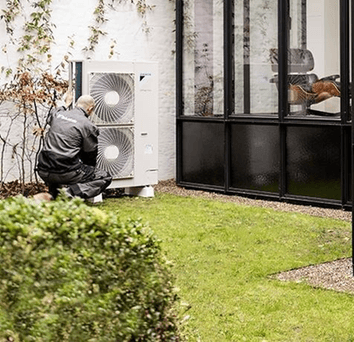 La photo d'une maison avec une pompe à chaleur, vue de l'extérieur, avec un technicien installateur qui travaille sur la pompe.